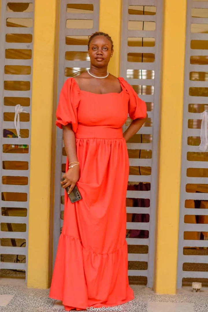 Grace standing in a coral dress outdoors against a warm yellow wall